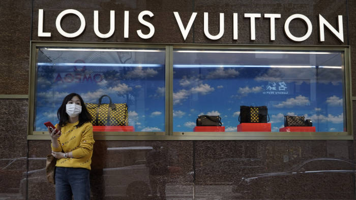A young woman stands outside a Louis Vuitton store in China wearing a face mask.